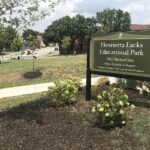 This picture features the sign of a park located in Baltimore that has been renamed to "Henrietta Lacks Educational Park". The sign is green and surrounded by other greenery. It features the name of the park and also the address.